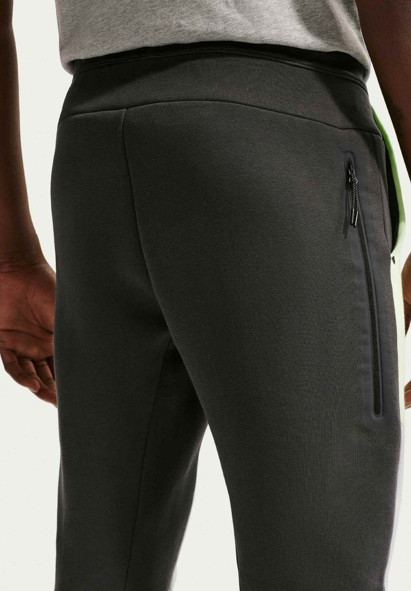 Nærbillede af person iført mørkegrå bukser med lynlåslomme i siden og en lys grå skjorte stoppet ind i taljen.