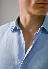 Vaaleansininen pellavapaita pehmeällä pinnalla, jossa on terävä kaulus ja kaksi nappia kaula-aukossa, paljastaen kangaspintaa.