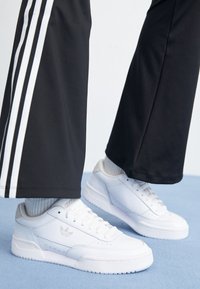 Witte leren sneakers met geperforeerde details en subtiele branding, gecombineerd met zwarte sportbroeken met witte zijkanten.