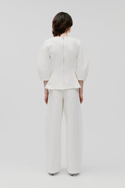 Vrouw met kort donker haar draagt een witte top met pofmouwen en een rits aan de achterkant en bijpassende broek met wijde pijpen tegen een effen lichtgrijze achtergrond.