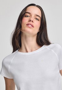 Vit kortärmad T-shirt med rund halsringning och en liten guldlogotyp på vänster bröst. Slätt tyg med en avslappnad passform.