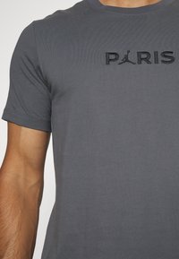 Grå t-shirt med broderad text "PARIS" och en sportlogotyp. Tyget har en slät struktur, och ärmarna är korta med en klassisk passform.