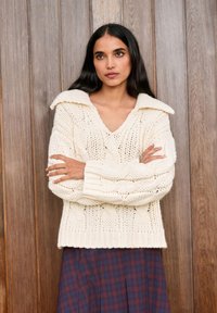 Krém színű, kötött mintás pulóver széles gallérral, V-nyakkal és bordázott mandzsettákkal, sötétkék és bordó árnyalatú kockás szoknyával párosítva.