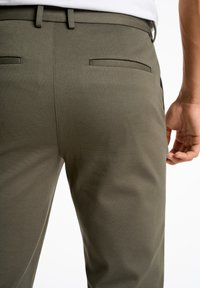 Pantalon vert olive avec une texture lisse, des poches arrière détaillées et une coupe ajustée, avec une ceinture et des passants de ceinture visibles.