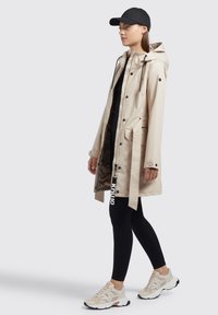 Beige vettore impermeabile con cappuccio, tasche frontali e coulisse. Indossato sopra leggins neri e sneakers bianche con accenti colorati.