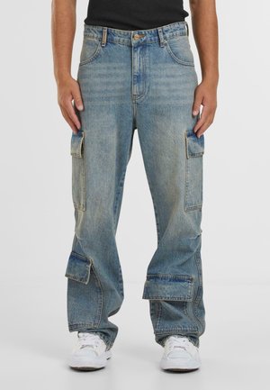 Pantaloni cargo in denim azzurro chiaro con più tasche con patta indossati da una persona con scarpe da ginnastica bianche e top nero, in piedi con le mani lungo i fianchi.