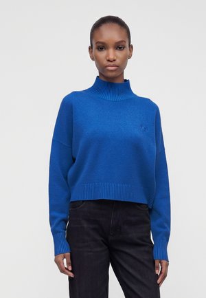 Mujer con un suéter de cuello alto acanalado de color azul brillante y pantalones negros, de pie frente a un fondo claro y sencillo.