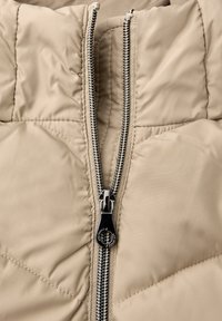 Veste matelassée beige avec col montant. Caractéristiques d'une double fermeture éclair noire, de détails de panneau cousus et d'une texture de tissu lisse et brillante.