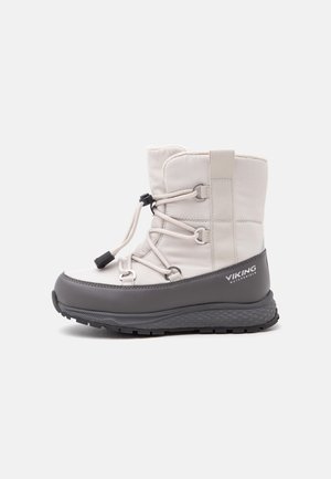Bota de invierno con parte superior beige claro, sección inferior gris impermeable, cordones frontales ajustables y suela negra texturizada para tracción.