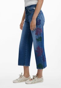 Jeans med vida ben i mellanblått med färgglad lövbrodyr på sidan. Matchat med ljusbeige snörskor.