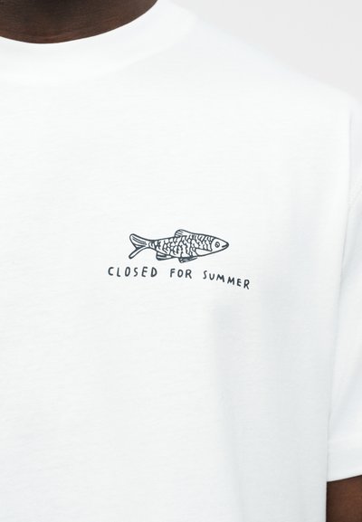 Chemise blanche avec une petite illustration noire de poisson et le texte "FERMÉ POUR L'ÉTÉ" imprimé en dessous sur la poitrine.