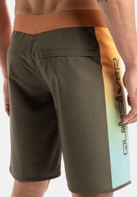 Bruna och olivgröna badshorts med en flerfärgad gradient på sidan, med en bakficka och en Quiksilver-logotyp.