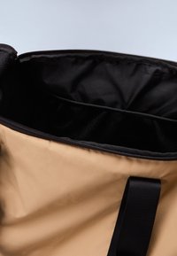 Tan fabric bag with a black interior, featuring a zip closure and a flat, webbed shoulder strap. Interior has a mesh compartment.