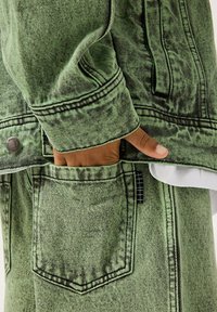 Giacca in denim verde con tessuto testurizzato, cuciture visibili e bottone in metallo, abbinata a pantaloni coordinati dotati di tasca e accento a motivo a griglia.