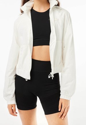 Vrouw met een wit jack met ritssluiting over een zwart sportbeha en zwarte biker shorts met hoge taille, staand met ontspannen armen.