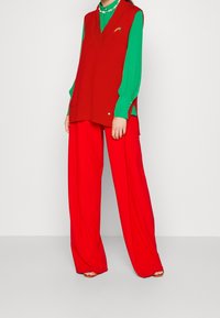 Veste rouge tricotée portée sur un chemisier vert, associée à un pantalon rouge à jambes larges et des sandales orange. Détails remarquables : un petit motif brodé.