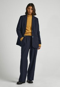 Blazer a rayas azul marino con pantalones de pierna ancha a juego, combinado con un cuello de tortuga mostaza. El blazer cuenta con solapas con muescas y cierre de botón.