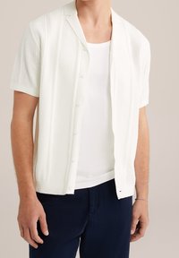 Vit kortärmad cardigan med texturerat stickmönster, knäppning fram, öppen krage, bärs över en vit linne, tillsammans med mörka byxor.