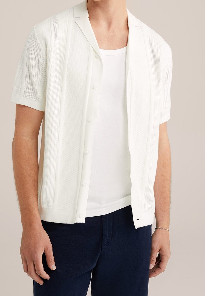 Vit kortärmad cardigan med texturerat stickmönster, knäppning fram, öppen krage, bärs över en vit linne, tillsammans med mörka byxor.