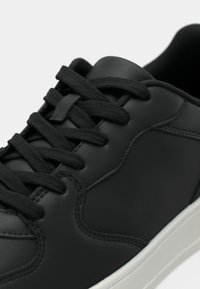 Artimas juodų odinių sportbačių, juodais raišteliais ir balta padais, vaizdas, kuriame matyti išsamios siuvimo linijos ir paminkštinta apykaklė.