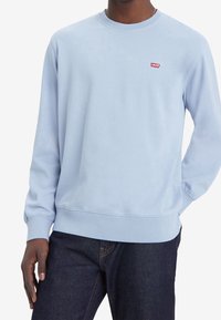 Sudadera azul claro con cuello redondo y puños acanalados. Presenta un pequeño logo rojo de Levi's en el pecho. Textura de tela suave.