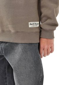 Barnet bär en taupe långärmad tröja med etiketten "Band of Rascals" och blekta svarta jeans, stående med handen avslappnat vid sidan.
