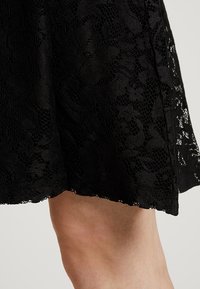 Jupe en dentelle noire présentant un motif floral, une surface texturée et un ourlet festonné. Le design est léger et semi-transparent.