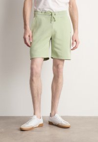 Homme portant un short vert clair à cordon et des baskets blanches, debout devant un fond clair uni.