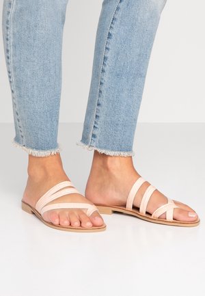 Des pieds portant des sandales plates à lanières rose clair associées à un jean cropped bleu clair à ourlet effiloché sur un sol blanc.