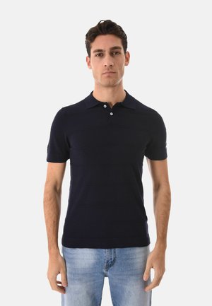 Uomo che indossa una maglietta polo nera a maniche corte e jeans azzurri chiari, in piedi davanti a uno sfondo bianco.