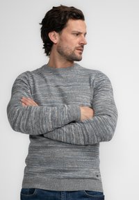 Grau gestrickter Pullover mit einem strukturierten Muster, das blaue und weiße Sprenkel aufweist. Rundhalsausschnitt und gerippte Bündchen, kombiniert mit dunklen Jeans.