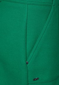 Grön fleece-tyg med en dragkedjeficka, framträdande sömmar och en märkesmetallaccent. Slät textur med en livlig färg.