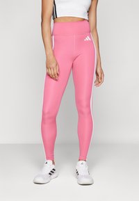 Roze high-waisted leggings van rekbare stof hebben witte zijstrepen en een Adidas-logo nabij de heup, gecombineerd met witte sportschoenen.