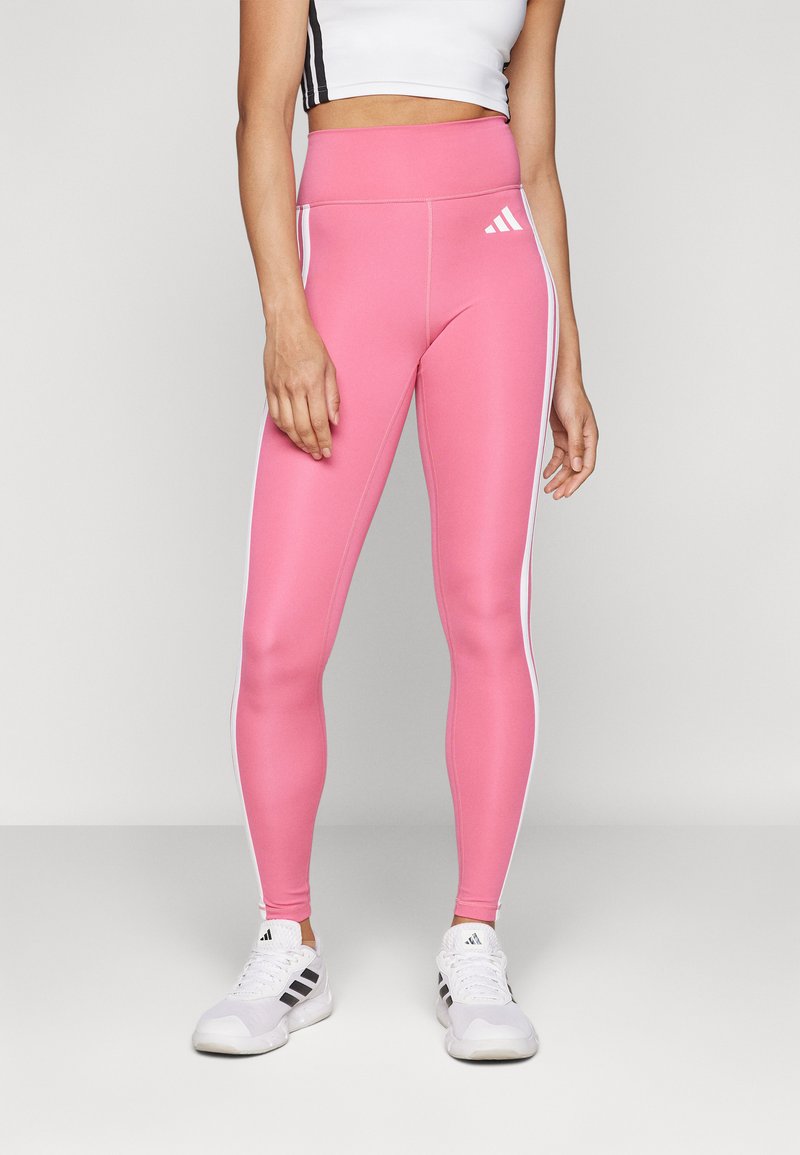 Roze high-waisted leggings van rekbare stof hebben witte zijstrepen en een Adidas-logo nabij de heup, gecombineerd met witte sportschoenen.