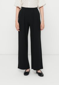 Pantalons larges noirs avec plis, une taille haute et une texture lisse. Associés à un chemisier à pois et des ballerines noires.