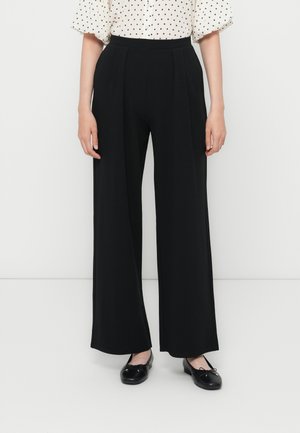Schwarze weit geschnittene Hosen mit Falten, einem hohen Bund und einer glatten Textur. Kombiniert mit einer gepunkteten Bluse und schwarzen Ballerinas.