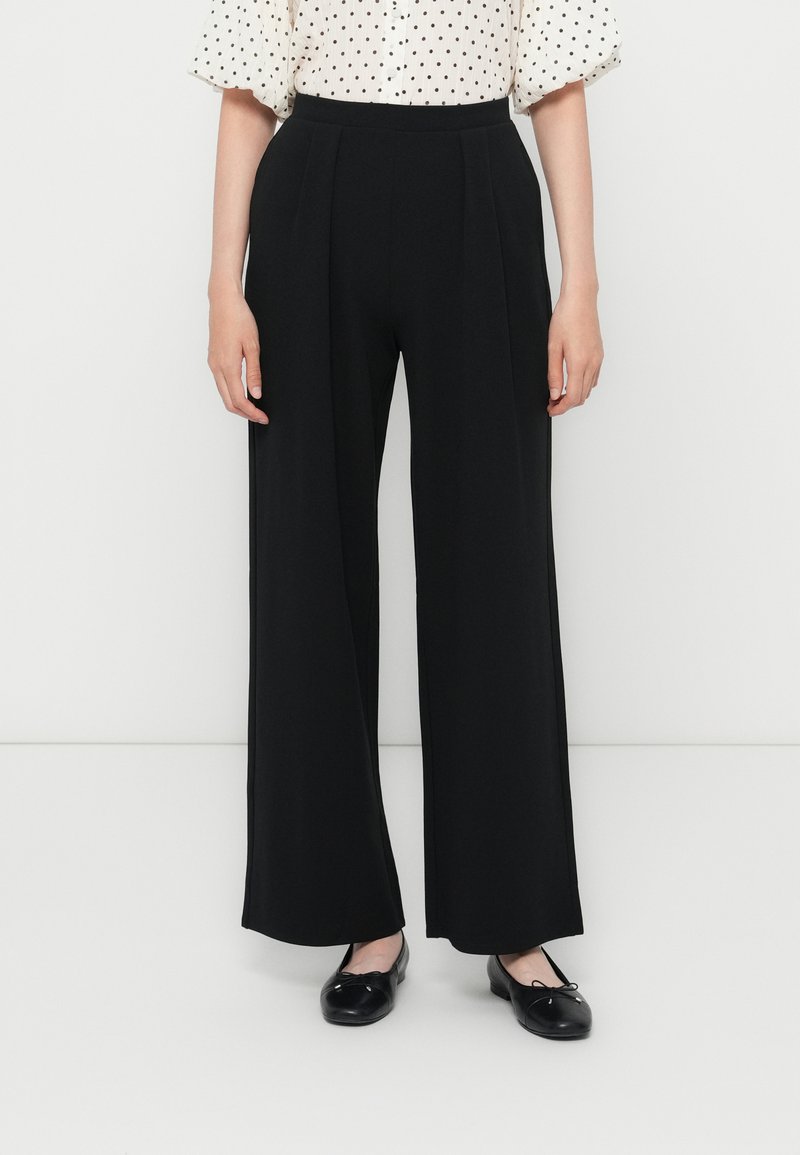 Pantalons larges noirs avec plis, une taille haute et une texture lisse. Associés à un chemisier à pois et des ballerines noires.