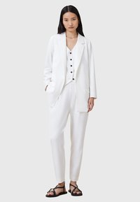 Completo bianco su misura composto da blazer, gilet con bottoni neri e pantaloni affusolati. Abbinato a sandali neri. Texture liscia, design minimal.
