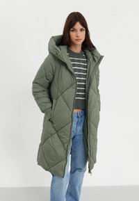 Parka longue matelassée en vert olive avec capuche. Dotée d'un motif diagonal et de poches latérales. Portée sur un pull rayé gris et un jean bleu.