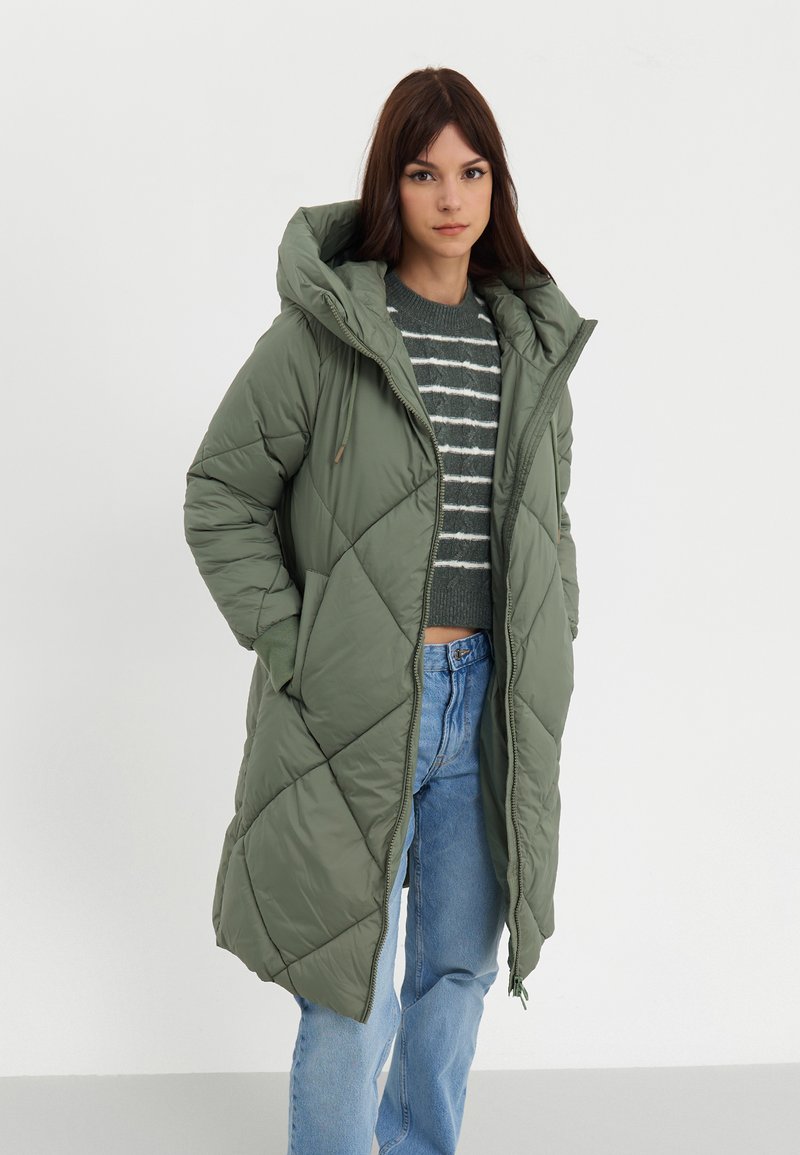 Parka longue matelassée en vert olive avec capuche. Dotée d'un motif diagonal et de poches latérales. Portée sur un pull rayé gris et un jean bleu.