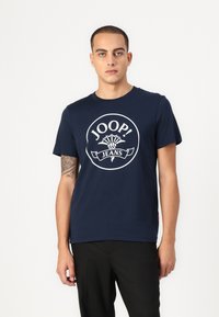 T-shirt en coton bleu marine avec un col rond, arborant un logo circulaire blanc qui dit "JOOP! JEANS" et un motif floral au-dessus.