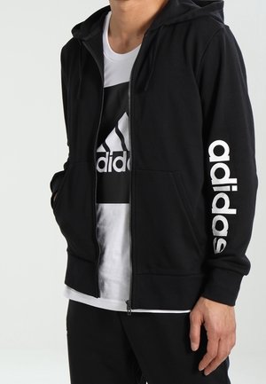 Schwarzer Kapuzenpullover mit Reißverschluss und weißem "adidas"-Schriftzug auf dem Ärmel sowie weißem T-Shirt darunter mit schwarzem Logo. Glatter Stoff.