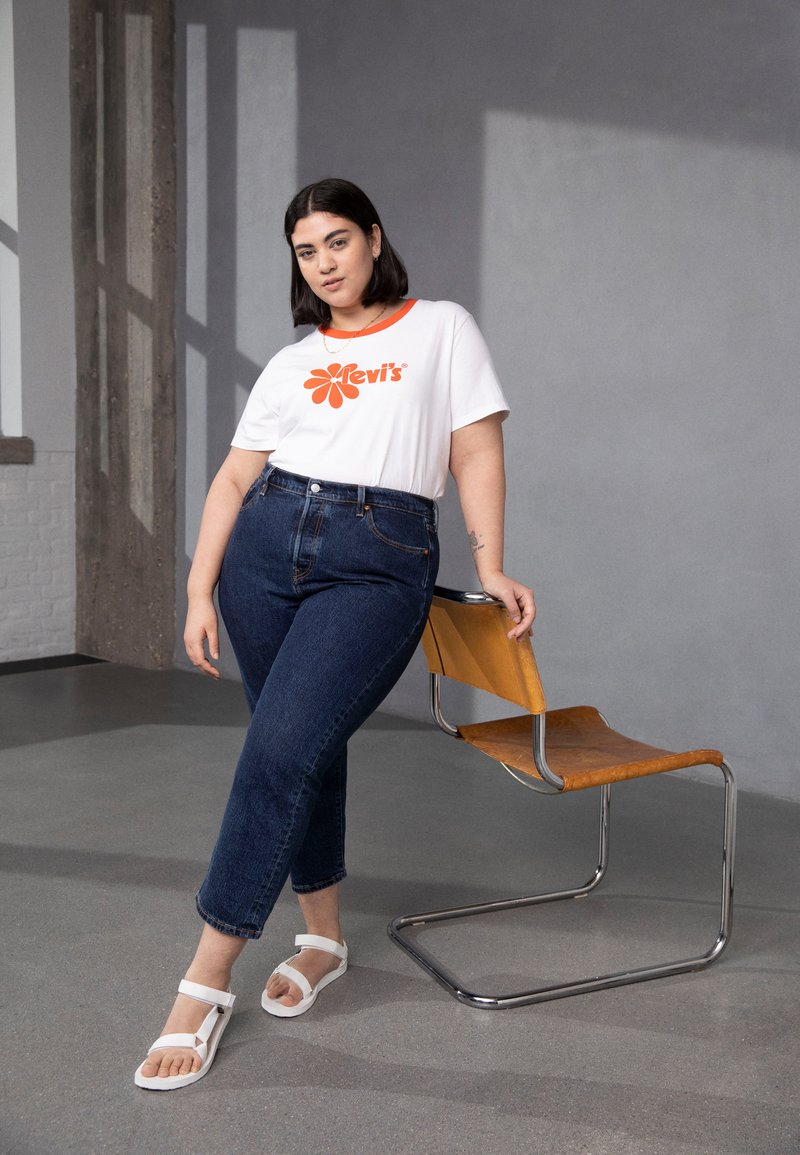 Eine Frau, die sich an einen braunen Lederstuhl lehnt, trägt ein weißes Levi's T-Shirt, dunkelblaue Jeans und weiße Sandalen und steht in einem minimalistischen Raum.