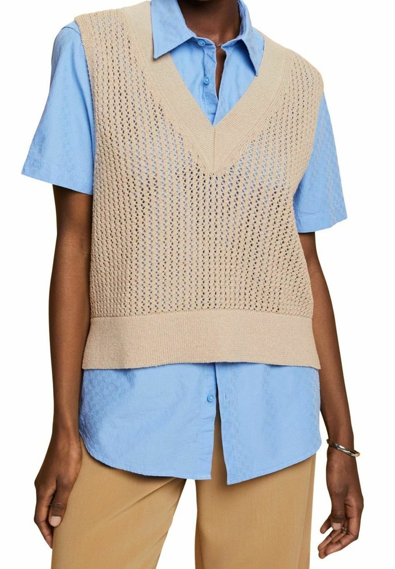 Personne portant un gilet sans manches en tricot beige par-dessus une chemise à manches courtes boutonnee bleu clair et un pantalon beige, avec un bracelet au poignet gauche.