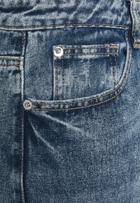Jeans de denim con un lavado azul y desgastado. El bolsillo trasero incluye un botón plateado y costuras amarillas contrastantes para mayor durabilidad.