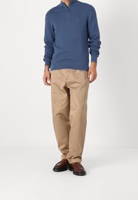 Mies, joka on pukeutunut siniseen vetoketjulliseen neulepuseroon, vaaleisiin housuihin ja tummanruskeisiin liukuisiin kenkiin, seisoo valkoista taustaa vasten.