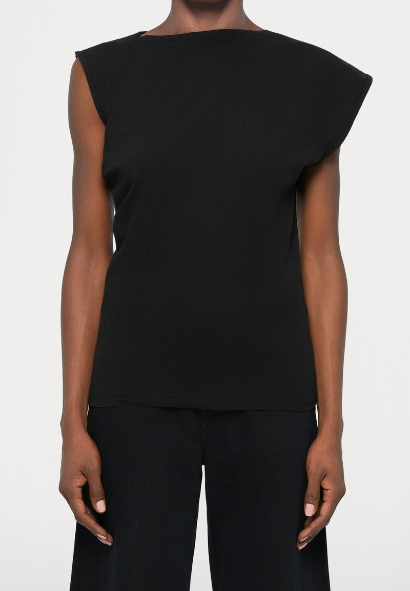 Personne portant un haut noir sans manches avec des épaules matelassées et un pantalon noir, debout devant un fond clair uni.