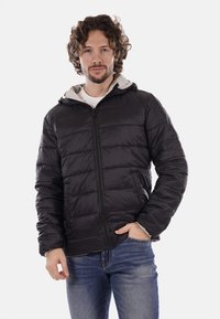 Chaqueta acolchada negra con capucha, con cremallera frontal y puños elásticos. Lleva puesta sobre una camisa de color claro, presentada sobre un fondo blanco.