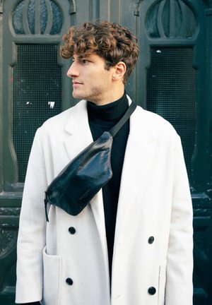 Schwarze Leder-Schultertasche mit glatter Textur, Reißverschluss und verstellbarem Riemen, getragen über einem weißen, zweireihigen Mantel.