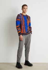 Strickpullover in Blau und Orange mit strukturierten Mustern; kombiniert mit grauen, schmal geschnittenen Hosen und schwarzen klobigen Schuhen. Einfache Hintergrund.